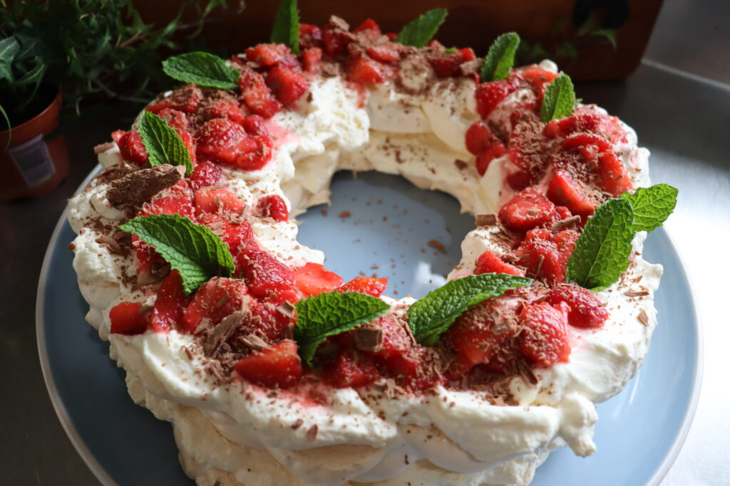 a pavlova wreath on blue plate topped with chocolate shavings, strawberries and mint leaves