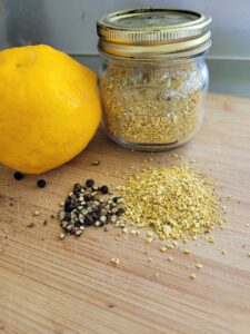 a small jar of lemon pepper seasoning sitting on a wooden board next to whole peppercorns and a lemon