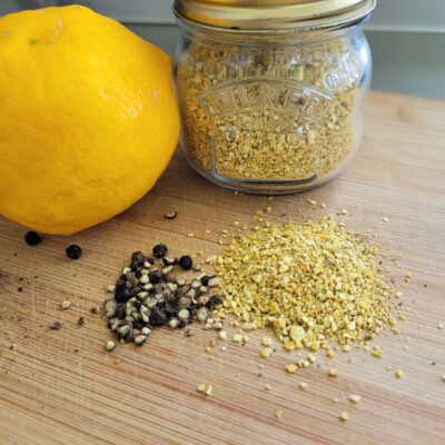 a small jar of lemon pepper seasoning sitting on a wooden board next to whole peppercorns and a lemon