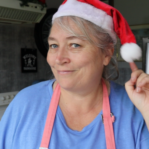 Farmers wife homestead standing in her kitchen with a Santa hat on and pointing to it with a smile on her face