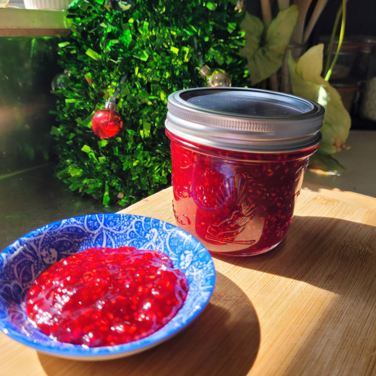 Homemade raspberry jam in a jar next to a small bowl of the jam on a wooden board