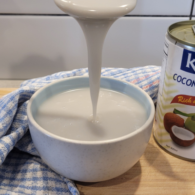 a bowl of dairy-free sweetened condensed milk on a wooden board with a can of coconut cream next to it. It is running off a spoon and is thick in consistency