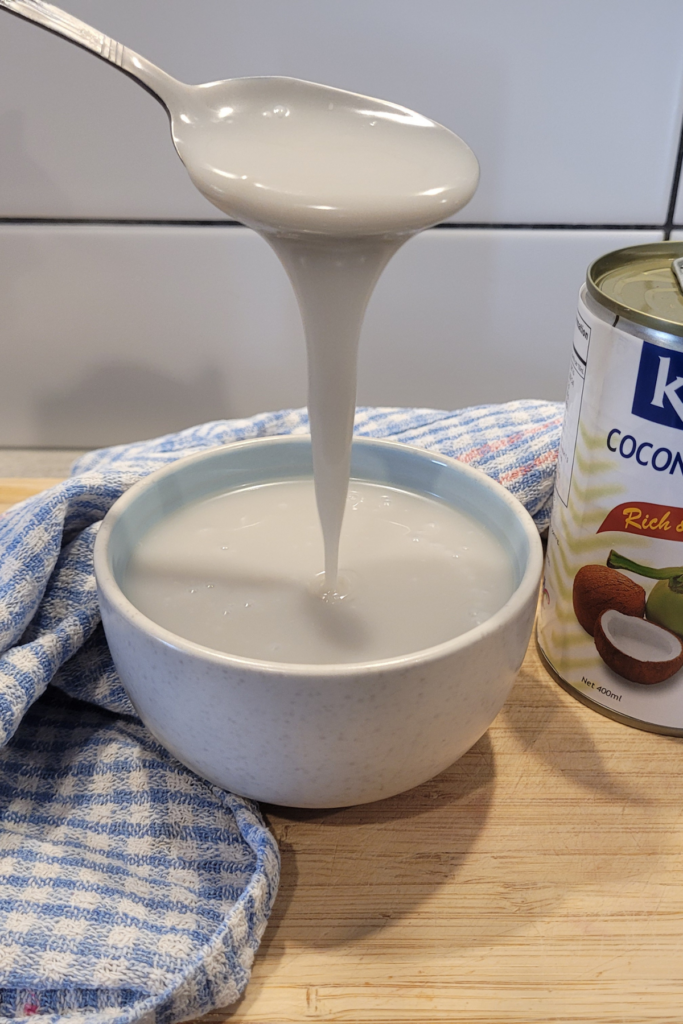 a bowl of dairy-free sweetened condensed milk on a wooden board with a can of coconut cream next to it. It is running off a spoon and is thick in consistency