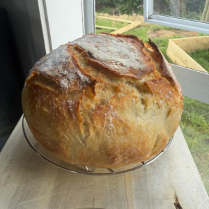 a beautiful round loaf of crusty bread sitting on a wire rack on a wooden board in front of a kitchen window