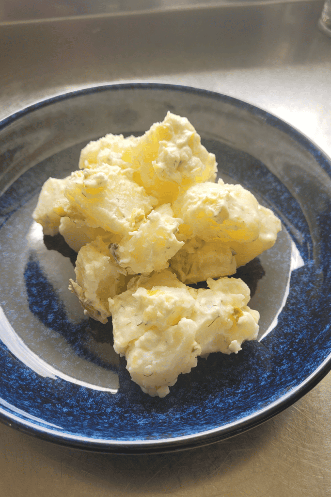 potato salad in a blue bowl on a wooden board it has a creamy dressing with gherkins in it