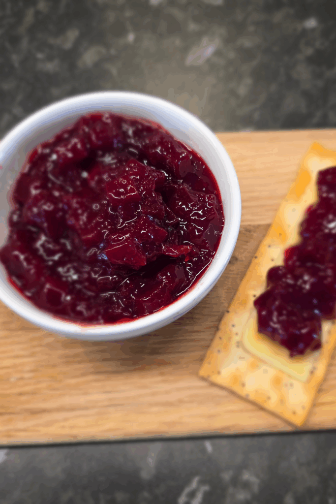Bowl of vibrant beetroot relish on a wooden board beside cheese and crackers.