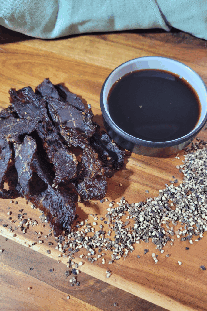 A square close-up of homemade beef jerky piled on a wooden serving board with peppercorns and soy sauce nearby. Soft natural light enhances the texture and deep colour of the jerky. Text reads “Homemade Beef Jerky | Simple Marinade, Big Flavour!”