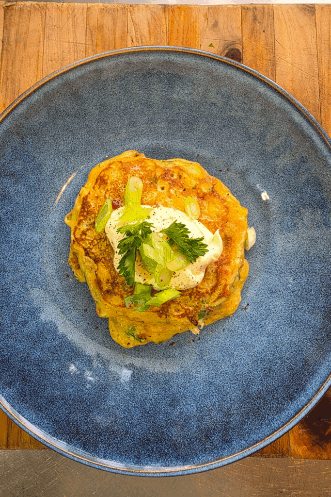Golden homemade corn fritters stacked on a blue plate topped with whipped sour cream and spring onions