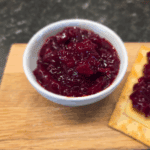 Bowl of vibrant beetroot relish on a wooden board beside cheese and crackers.