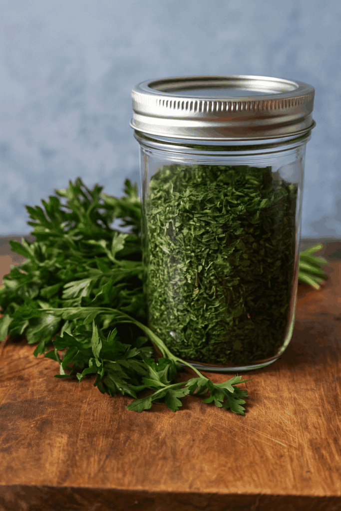 Glass jar of dried parsley beside fresh parsley on a wooden board, rustic blue background.