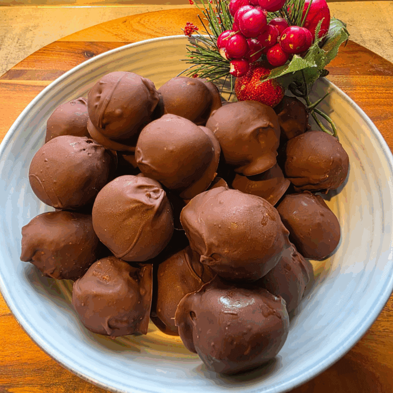 A bowl filled with glossy, chocolate-coated Mint Slice Choc Bombs, arranged on a wooden board with festive berries and greenery beside them.