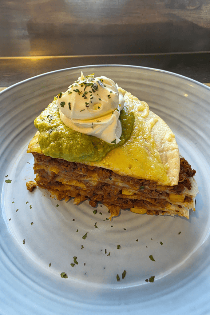 “Stacked Mexican-style tortilla bake layered with seasoned beef, cheese, and tortillas, sliced into wedges and topped with avocado and sour cream.”