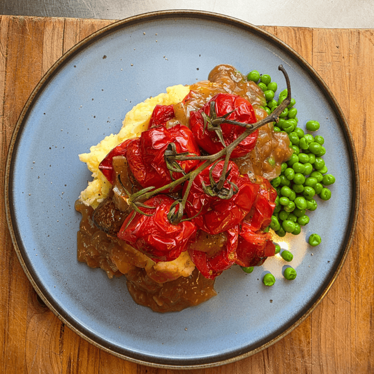 Plate of easy bangers and mash topped with rich onion gravy, roasted vine tomatoes & peas